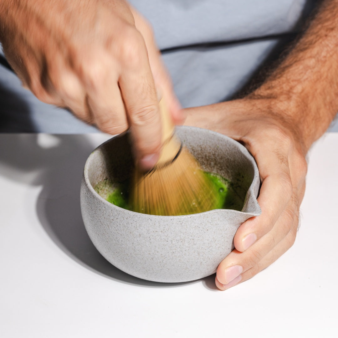 A close-up of hands whisking vibrant green Dakys Performance Matcha in a ceramic bowl using a traditional Matcha Whisk bamboo (chasen), creating a frothy, smooth texture. Ceremonial-grade Matcha whisked to create smooth foam, enhancing focus and mental clarity with every sip.