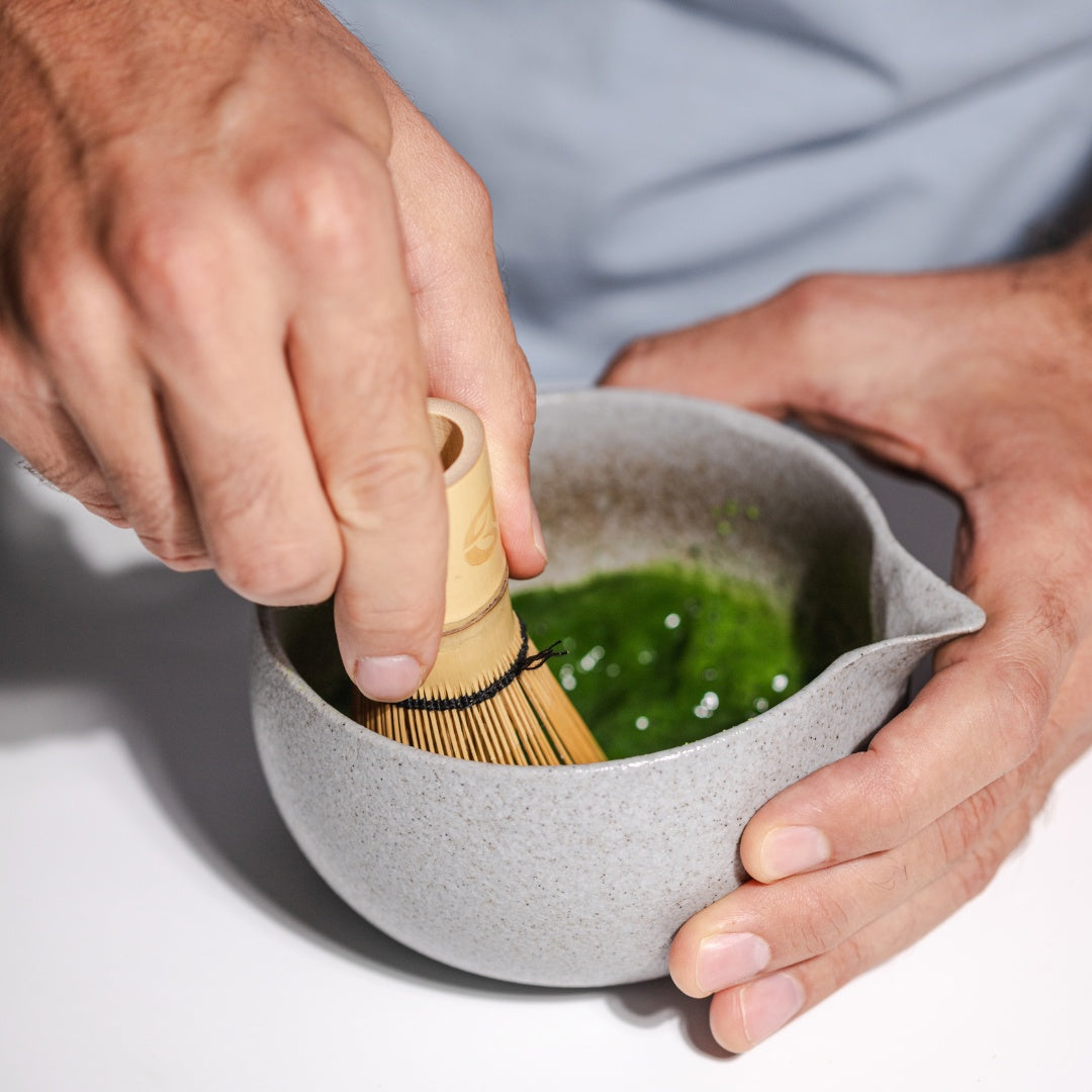 Hand whisking vibrant green matcha with a bamboo whisk in a ceramic bowl.