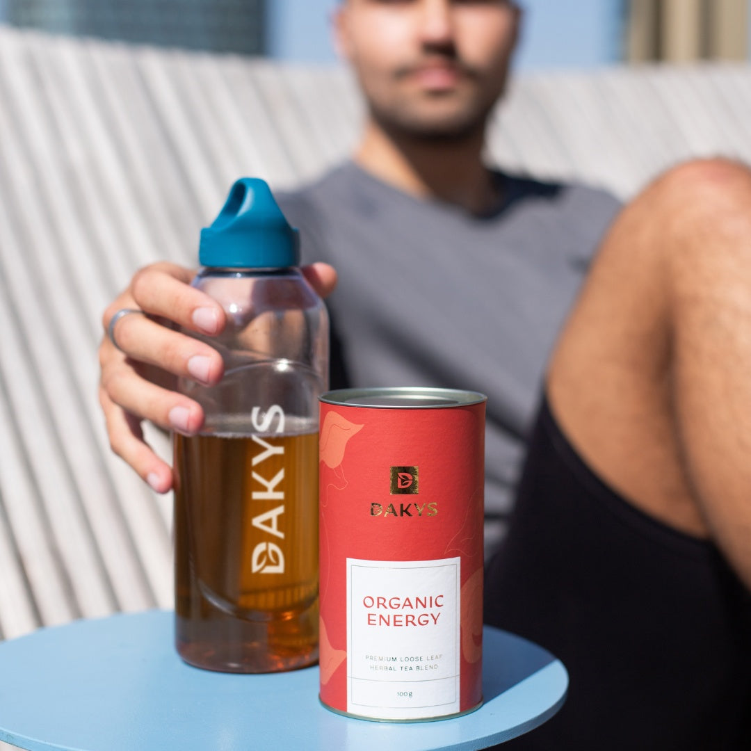 Man relaxing outdoors with a bottle of Dakys performance drink and a canister of Organic Energy on a small table.