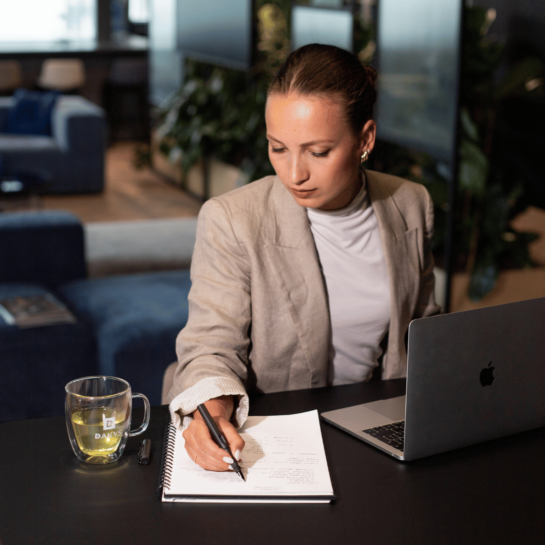 Focused woman writing in notebook with a glass of Dakys Gyokuro on the table, working in a modern office environment.
