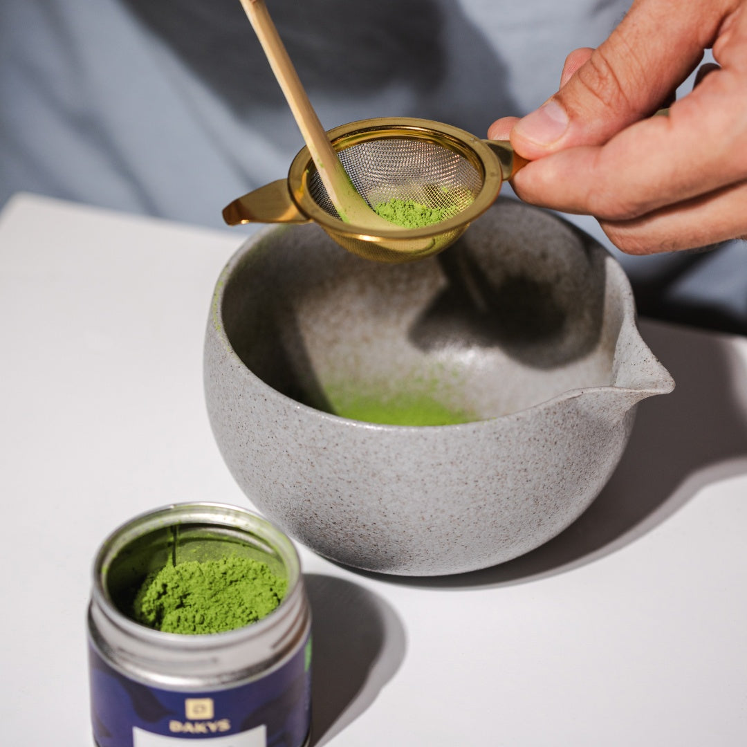 Close-up of Dakys Matcha being sifted through a fine strainer into a ceramic bowl, using a bamboo spoon. A jar of vibrant green matcha powder is open on the table, ready for preparation.
